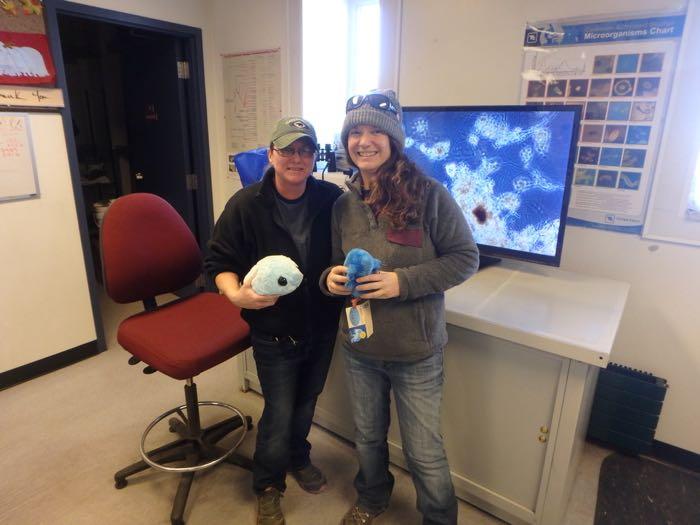 Two women with stuffed water bears standing in front of a monitor showing the tiny creatures that breakdown human waste. 