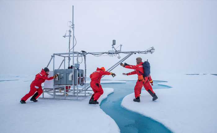 Three people push a sled of equipment across the ice