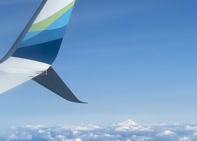 An image of the sky out of a plane window. Far in the distance a snow covered mountain peak can be seen. 