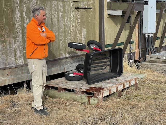 Dennis stands next to a wood palette with a wheelbarrow on it. On the corner of the wood palette is a puddle of fake blood. 