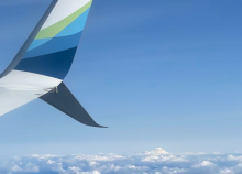 An image of the sky out of a plane window. Far in the distance a snow covered mountain peak can be seen. 