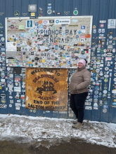 Liza stands in front of a blue wall covered in stickers. A white sign displays "Prudhoe bay" and a yellow sign displays "Deadhorse"