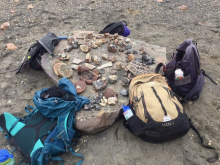 A bunch of small rocks sit on a boulder surrounded by backpacks. 