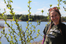 A woman with brown hair in braids in a green t-shirt stands in front of some foliage and a lake. 