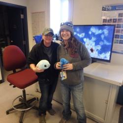 Two women with stuffed water bears standing in front of a monitor showing the tiny creatures that breakdown human waste. 