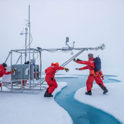 Three people push a sled of equipment across the ice